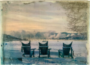 Four wooden chairs facing snowy landscape with steam, hills, boats, under cloudy sky.